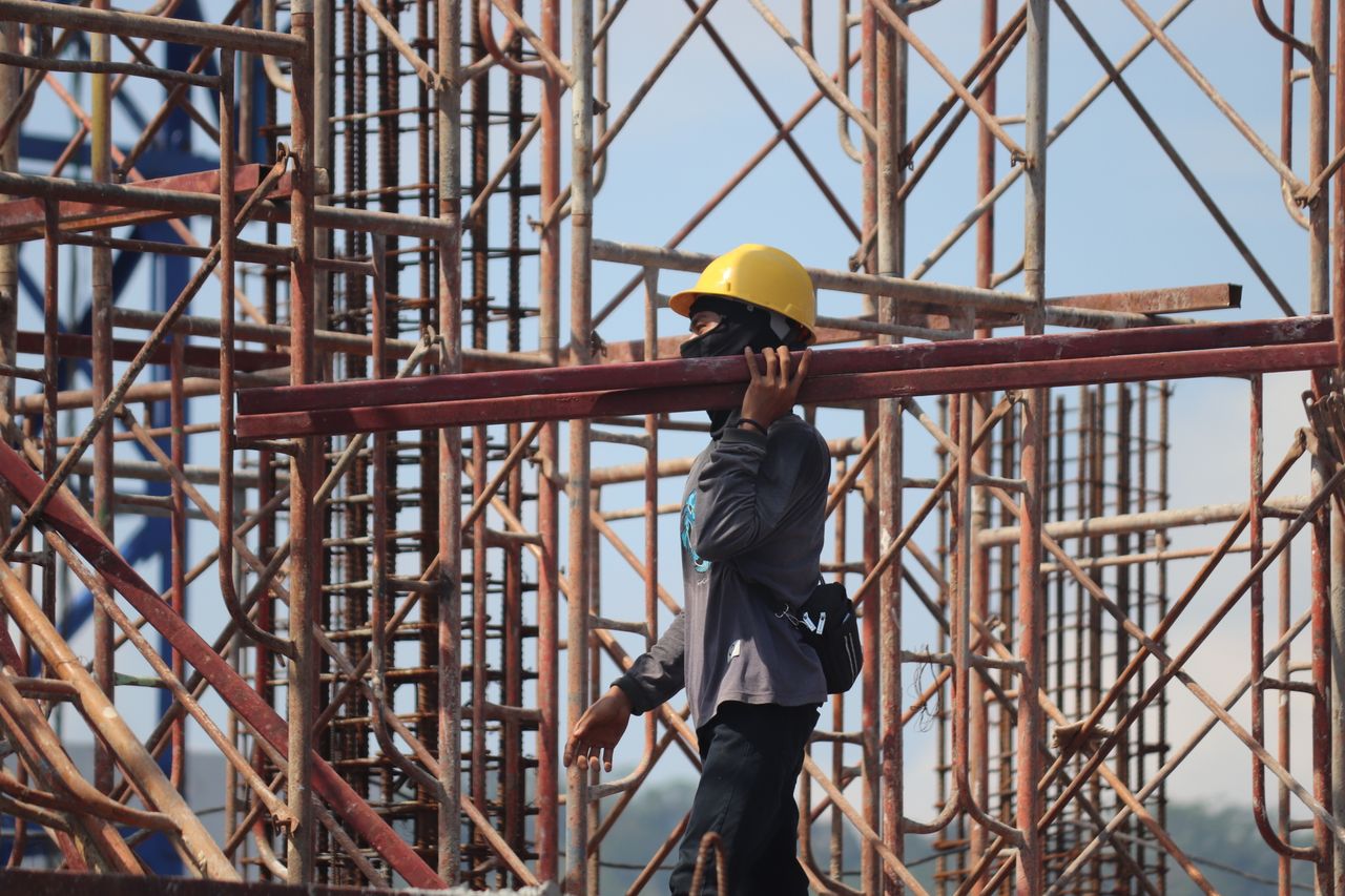 A construction worker with safety gear carrying a metal beam amidst scaffolding at a busy construction site.